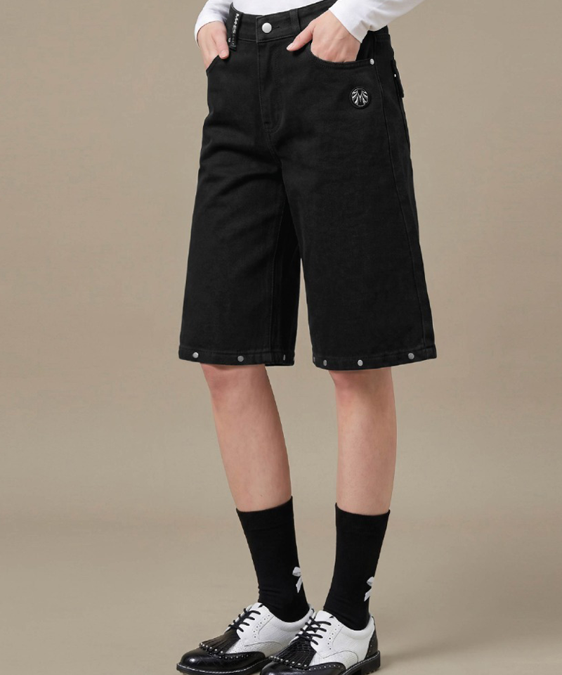 A person wearing black denim shorts with silver rivet details, styled with a white long-sleeve top, black socks, and black-and-white oxford shoes against a neutral beige background.