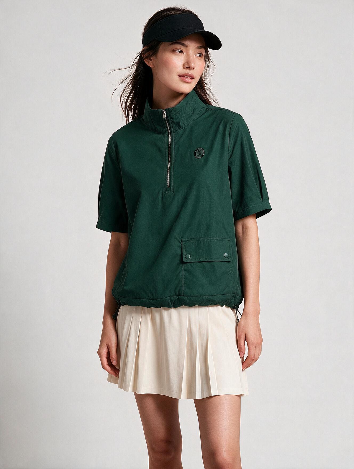 A woman modeling a dark green short-sleeve quarter-zip pullover with a small circular chest logo, paired with a cream pleated mini skirt and a black baseball cap, against a light gray studio background.